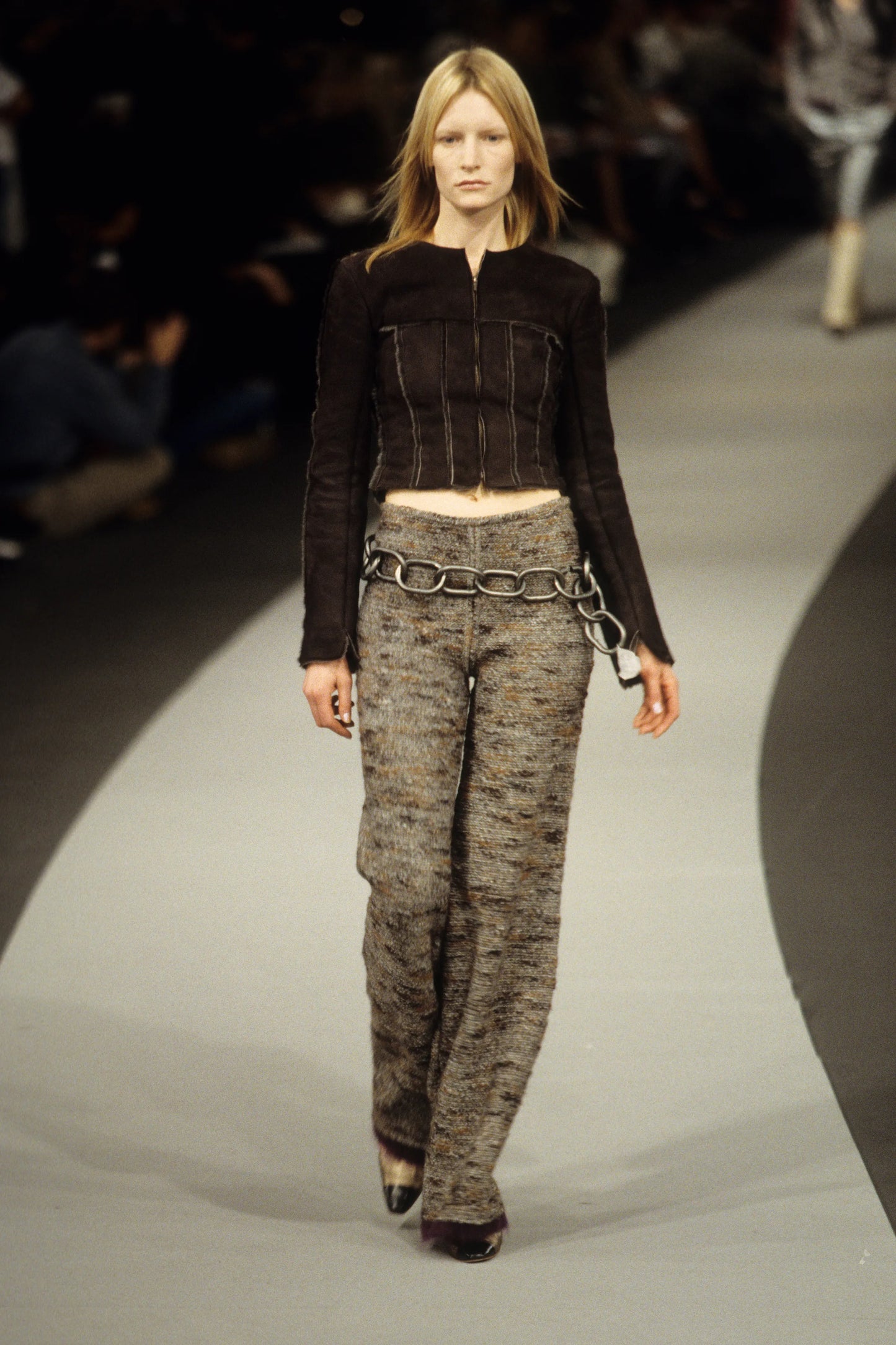 Model walking on a runway wearing a black top and patterned pants with chain details.