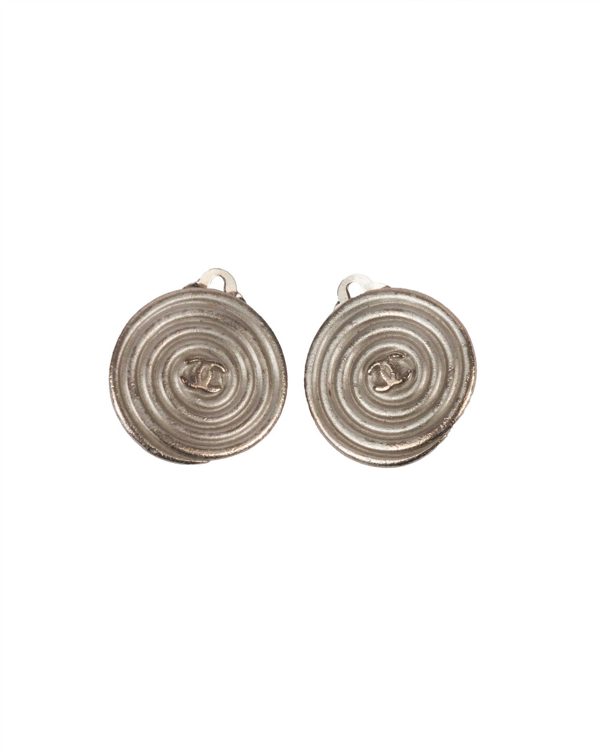 A pair of round silver-tone metal clip-on earrings with a spiral relief design and central CC logo.