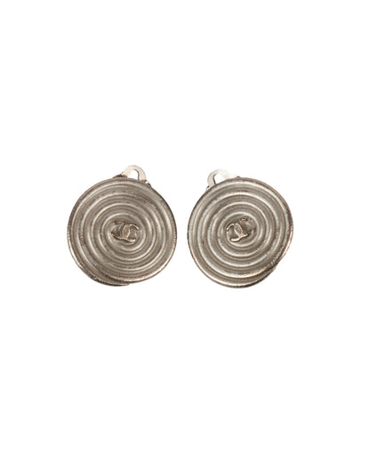 A pair of round silver-tone metal clip-on earrings with a spiral relief design and central CC logo.