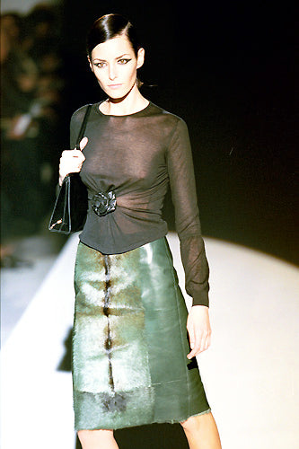 A model walks a fashion runway wearing a sheer black long-sleeve top with a sculptural waist detail and a green textured midi skirt, holding a black handbag.