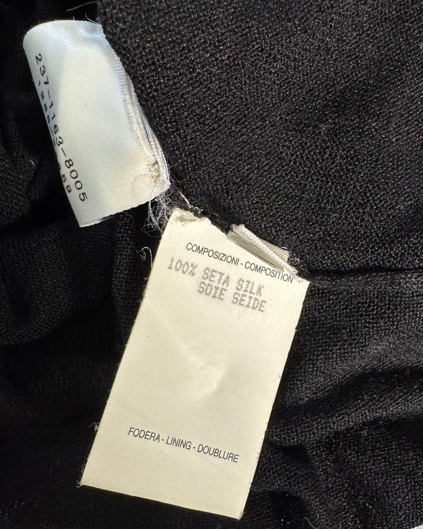 Close-up of a black fabric garment showing two tags: a white sewn-in size label and a larger white care/information tag reading '100% Seta Silk' and 'Foeda - Lining - Doublure'.