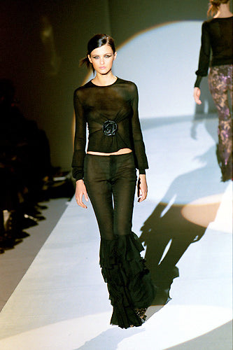 A model walks down a fashion runway wearing a black sheer long-sleeve top with a ruffled waist detail and matching black ruffled high-waisted pants.