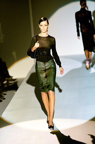 A model walks a fashion runway wearing a sheer black long-sleeve top and a shiny dark green pencil skirt, holding a black handbag.