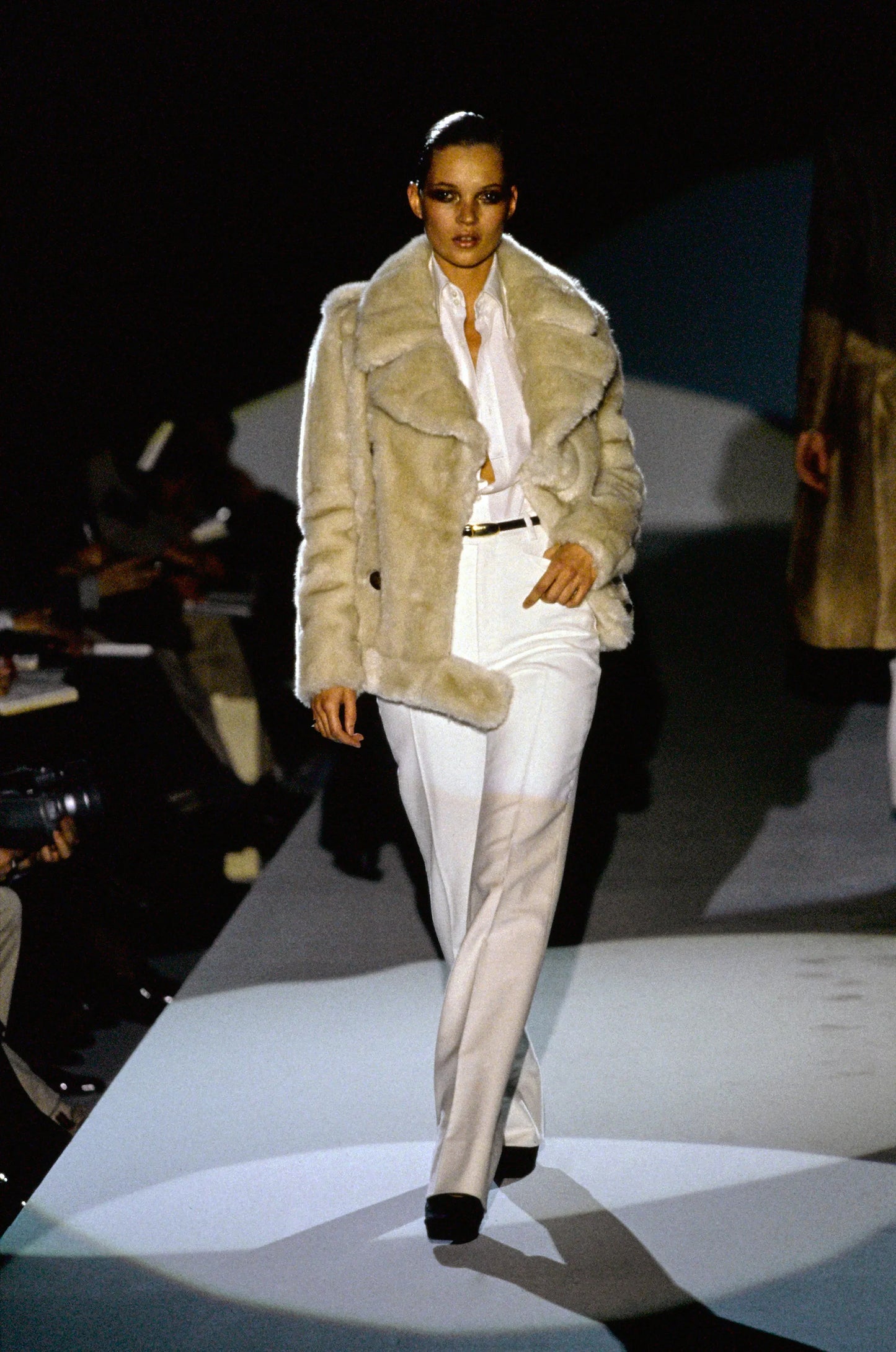 Model walking on a runway wearing a fur coat and white pants.