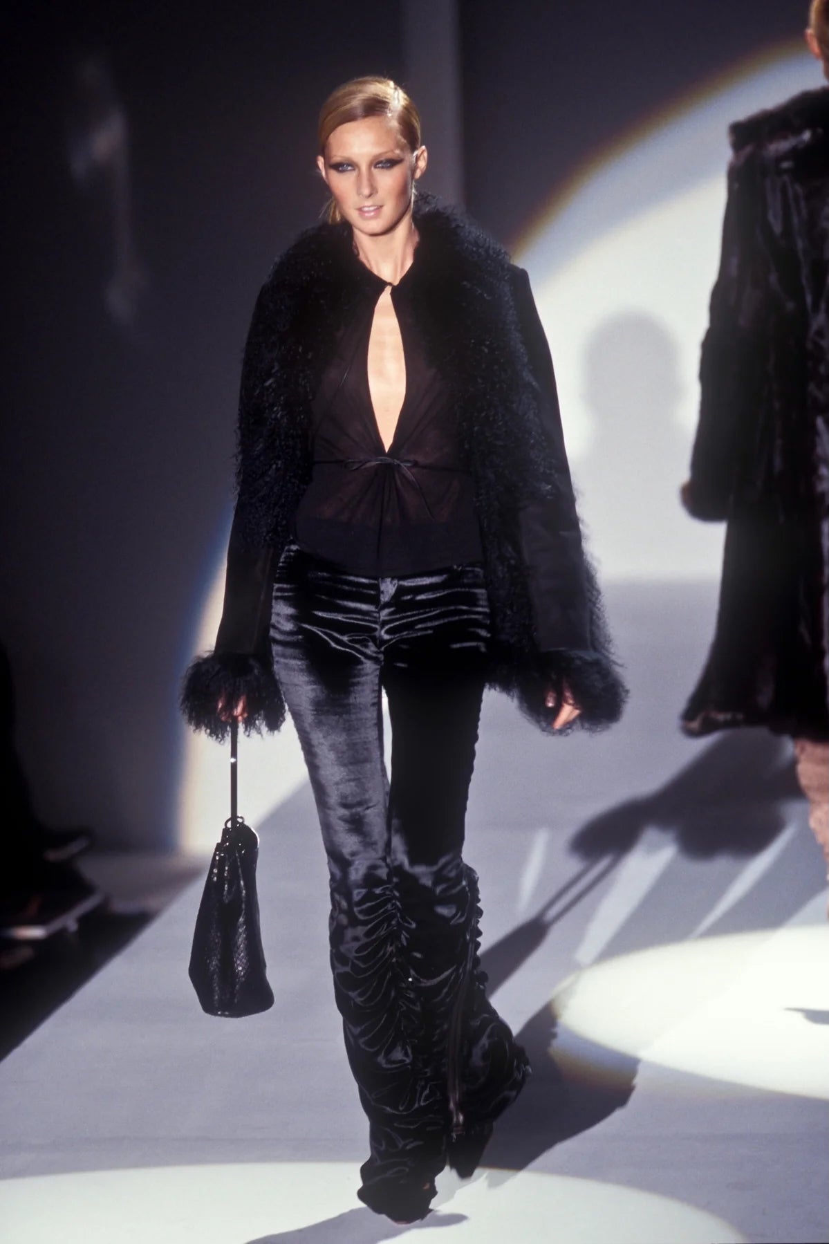 Model walking on a runway wearing a black fur coat, black top, and black pants with high boots.