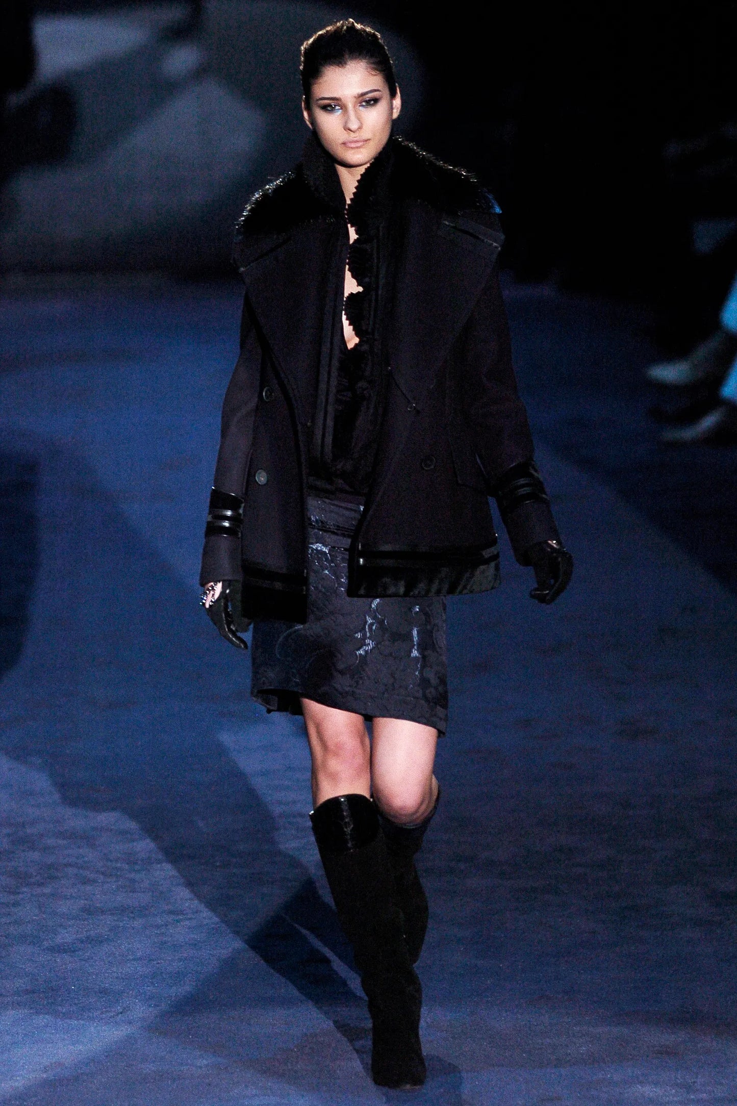 Model walking on a runway wearing a black coat, dress, and boots.
