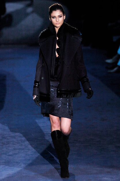 Model walking on a runway wearing a black coat, dress, and boots.