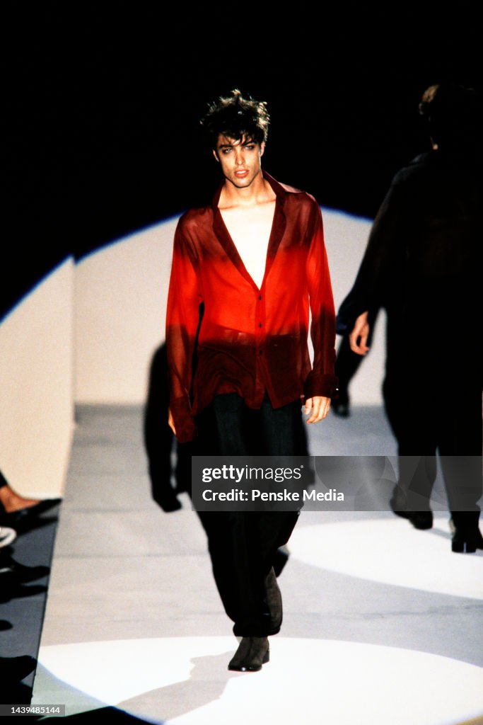 Model walking on a runway wearing a red jacket and black pants.