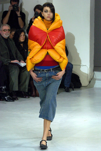 Model wearing a colorful puffer jacket and jeans on a runway with an audience.