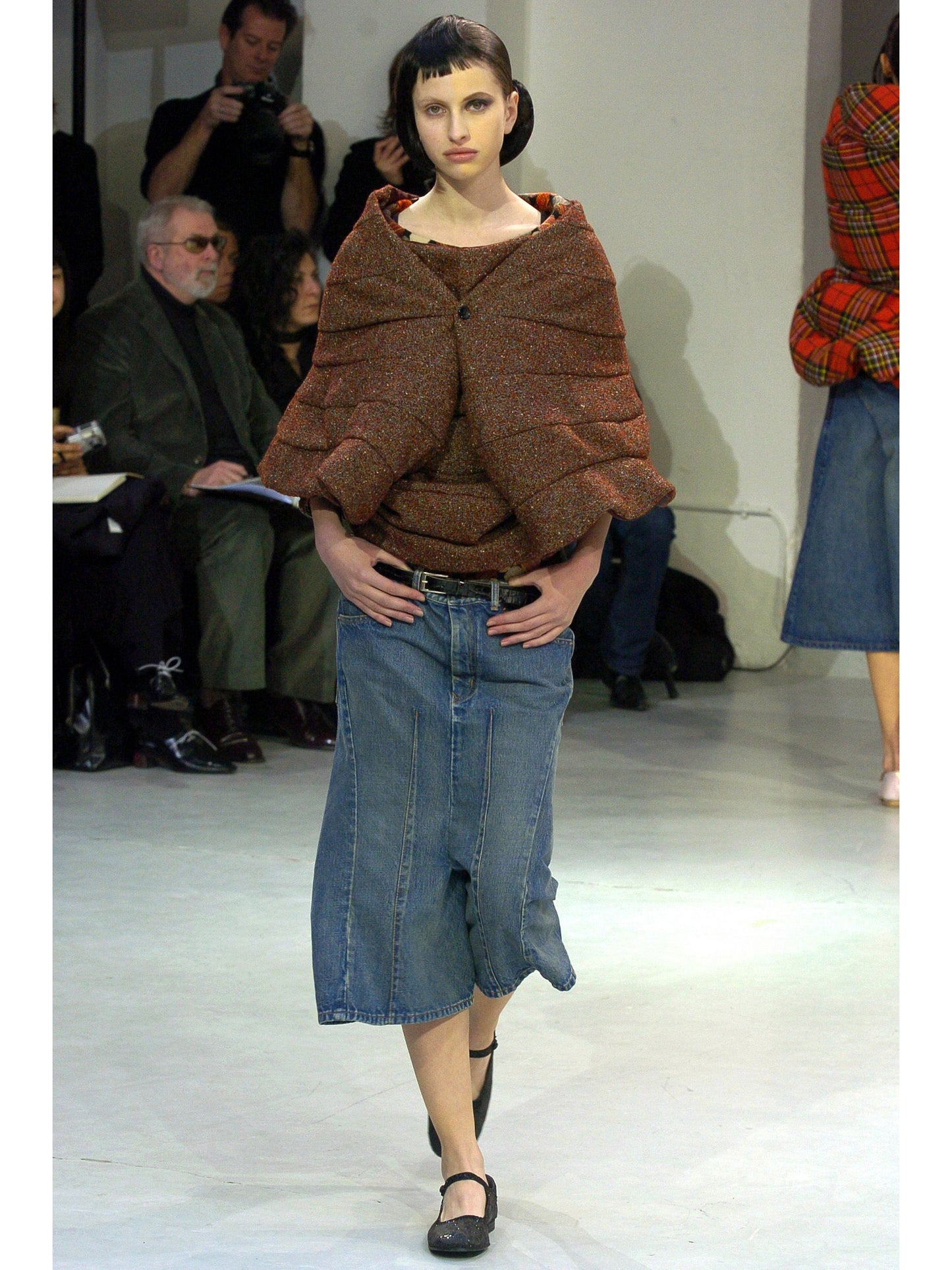 Model wearing a brown textured top and blue denim skirt on a runway with an audience.