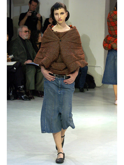 Model wearing a brown textured top and blue denim skirt on a runway with an audience.