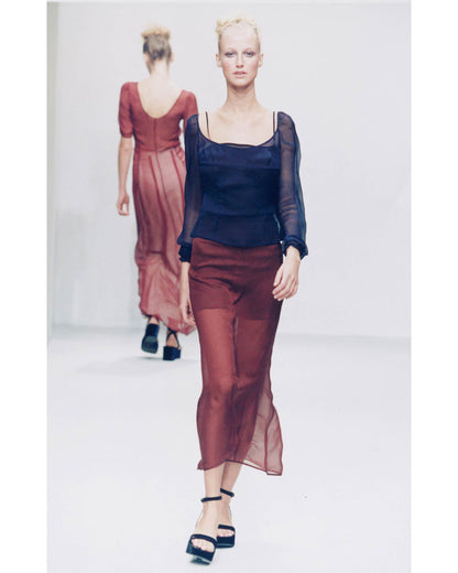 A runway fashion photo showing a model walking forward in a sheer dark navy top with long sleeves and a matching sheer rust-brown midi skirt. Another model is visible in the background wearing a rust-brown dress.