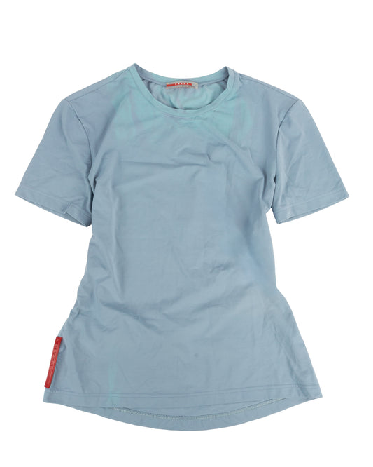 A powder blue short-sleeved technical t-shirt with a small zip pocket on the lower back and a red rubber Prada tag at the left hip.