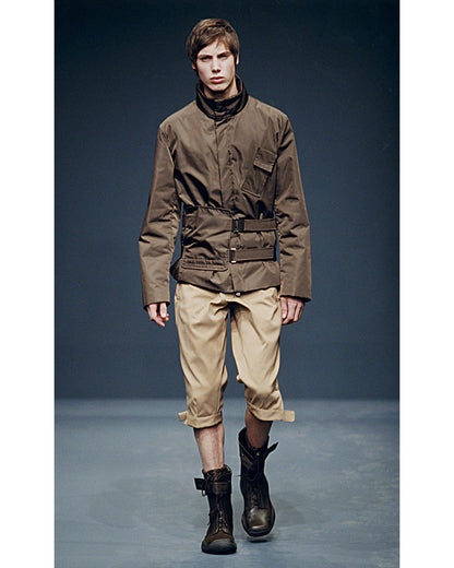 Model wearing a brown jacket and beige pants on a runway with a dark background