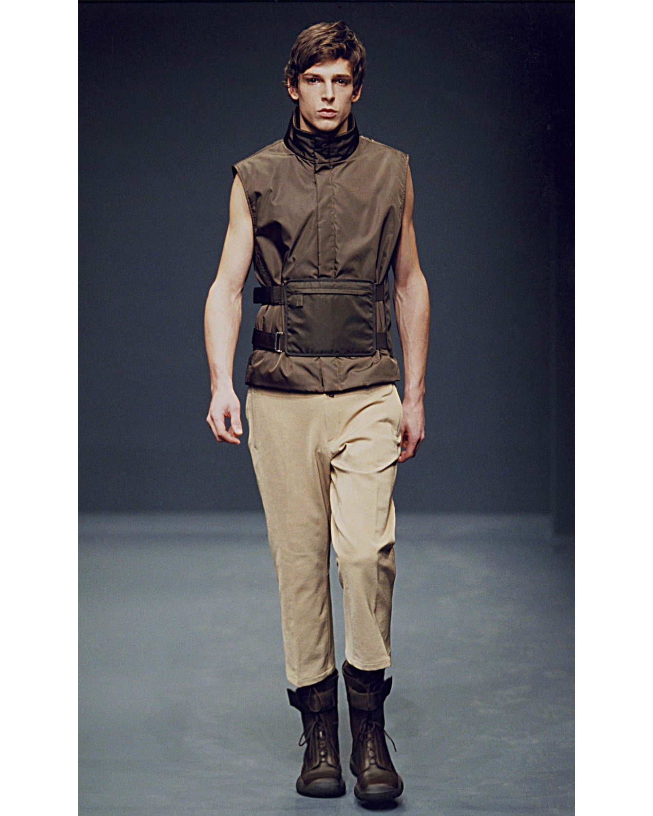 Man wearing a brown vest, beige pants, and dark boots on a runway.
