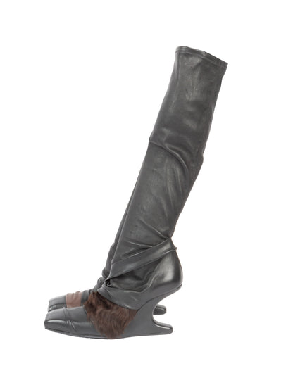 A black leather knee-high boot with a fur accent on the side, featuring an architectural wedge heel and a square toe.