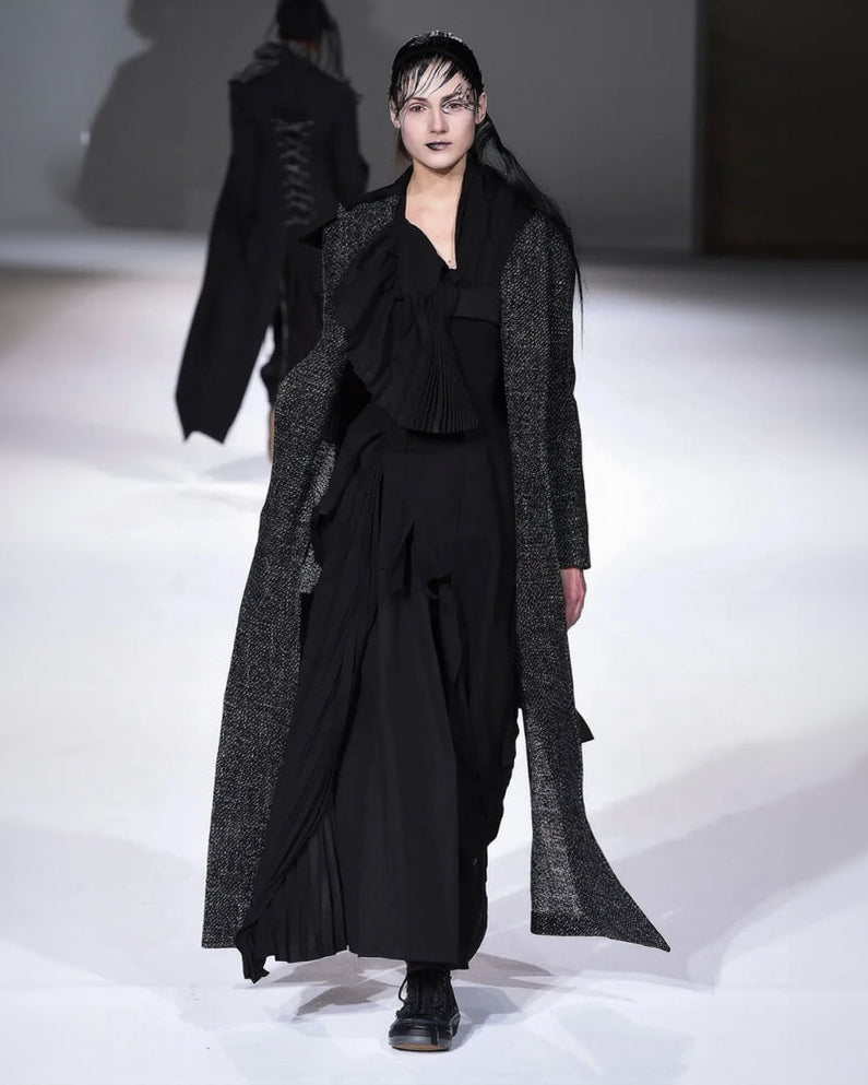Model wearing a black outfit with a long coat on a runway.